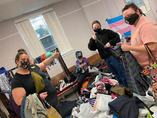 A group of individuals wearing masks stand and sit around a pile of clothes.  Some are holding clothing in their hands. | QueerDoc inclusive spaces
