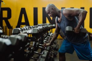 Dark-skinned masculine person with buzzed grey and black hair lifts a large dumbbell with their left arm. They are leaned forward with the left leg bent and left arm bracing on a rack of dumbbells. A yellow wall with large black letters is behind him partially spelling out 