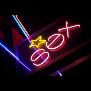 Sex in pink neon letters on a black background. There is also a yellow neon star, and neon tubing in blue, aqua, and pink,