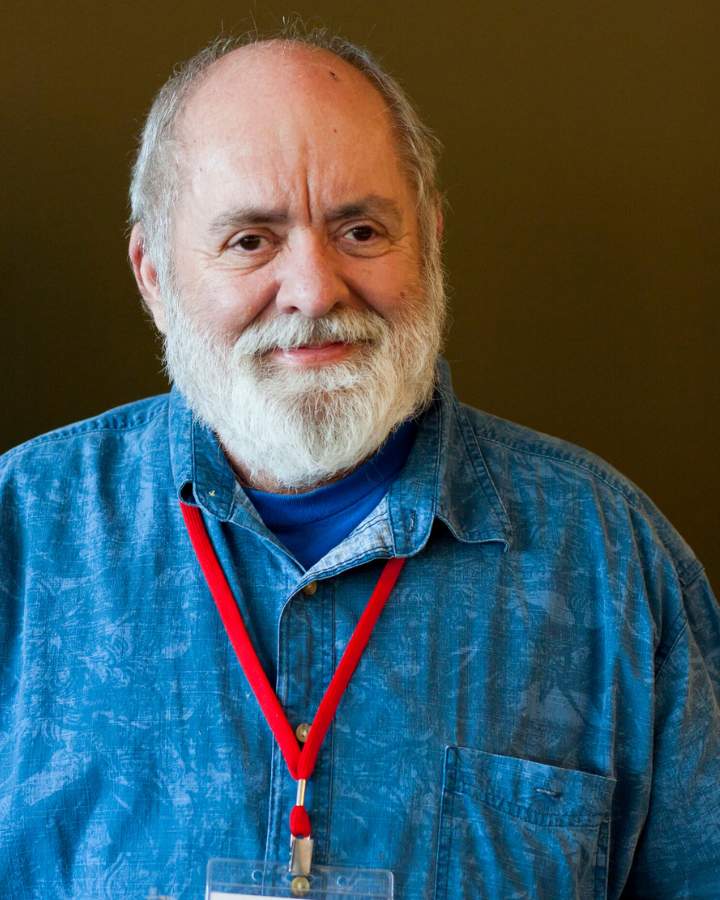 Jude Patton is a white man with a full white beard and mustache and short hair with a balding crown and dark eyes. He stands in front of a brown background, and wears a pattered blue button down shirt over a darker blue shirt. There is a red lanyard around his neck.