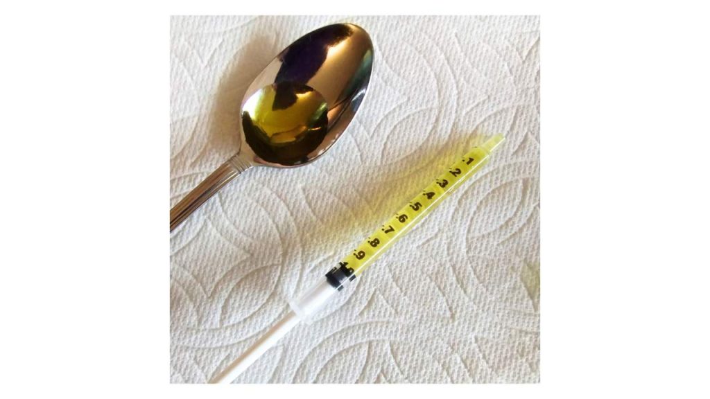 A stainless steel spoon with 1mL of olive oil in the bowl lies next to a 1ml syringe containing 1mL of olive oil.  Both are on top of a white paper towel. | QueerDoc syringes
